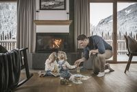 Vater spielt mit zwei Kindern vor Kamin in gemütlichem Wohnzimmer mit verschneiter Bergkulisse