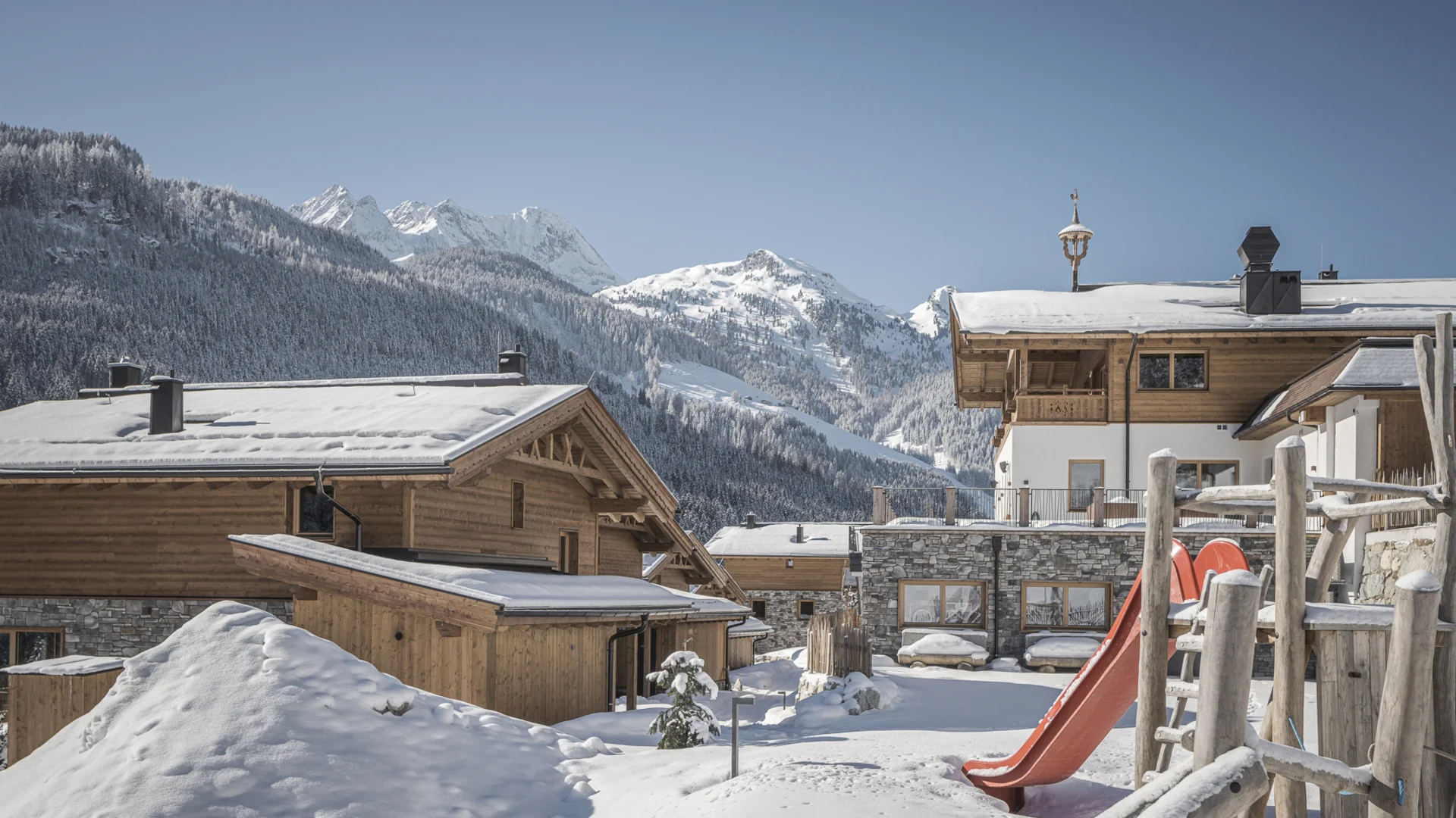 Snow-covered mountain cabins with playground and slide in front of snowy mountains